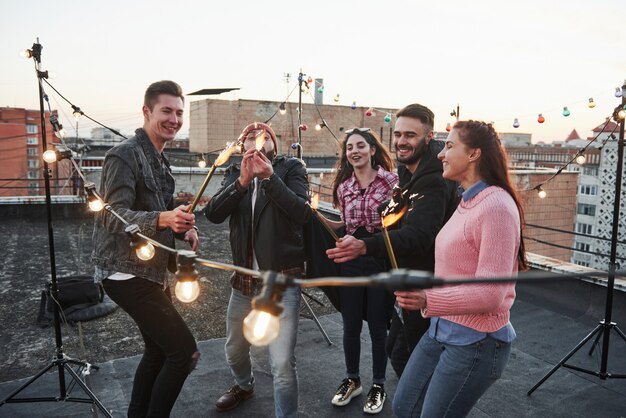 Guy in the red hat imagines that he is a dragon by tying to make flame bigger. Playing with sparklers on the rooftop. Group of young beautiful friends