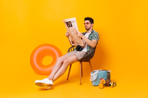 Free photo guy sits and reads newspaper on space of suitcase, pineapple and rubber ring.