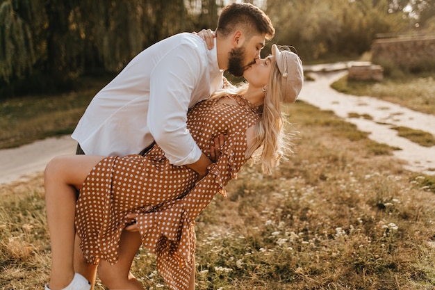 Guy in white shirt inclining his girlfriend during gentle and passionate kiss. Couple embracing amid park.