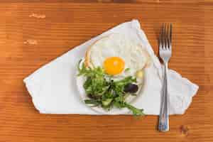 Free photo half fried egg with salad on plate and fork over the white napkin against wooden textured backdrop