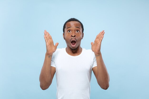 Half-length close up portrait of young african-american male model in white shirt on blue space. Human emotions, facial expression, ad or betting concept