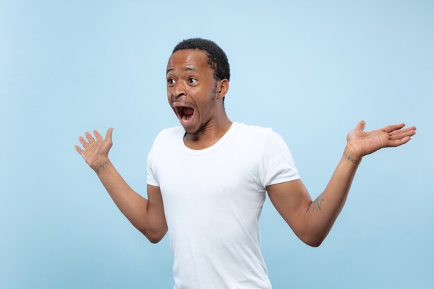 Half-length close up portrait of young african-american man in white shirt on blue background. Human emotions, facial expression, ad, sales concept. Inviting, looks shocked and astonished. Copyspace.