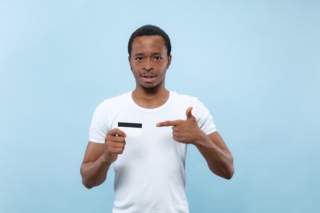 Half-length close up portrait of young african-american man in white shirt on blue background. Human emotions, facial expression, ad, sales concept. Pointing on the card. Payments, finance, bill.