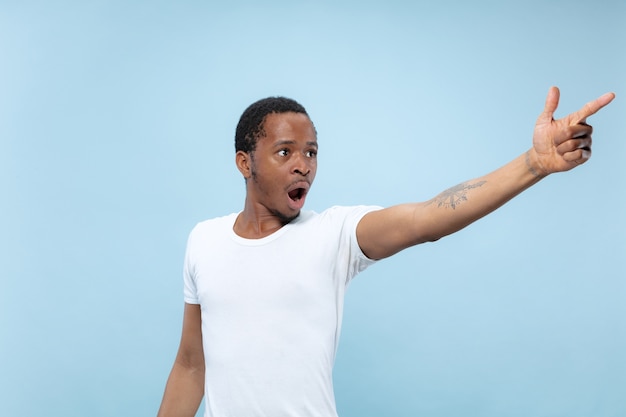 Half-length close up portrait of young african-american man in white shirt on blue background. Human emotions, facial expression, ad, sales concept. Pointing, choosing, astonished. Copyspace.