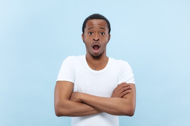 Half-length close up portrait of young african-american man in white shirt on blue background. Human emotions, facial expression, ad, sales, concept. Standing hands crossed, shocked and astonished.