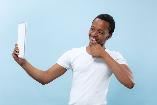 Half-length close up portrait of young african-american man in white shirt on blue background. Human emotions, facial expression, ad, sales, concept. Using tablet for selfie, vlog, talking.