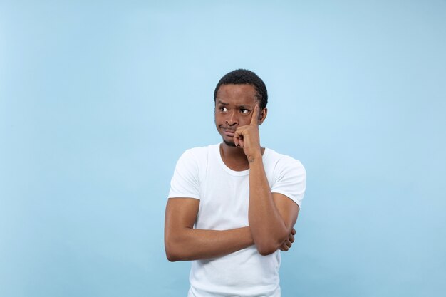 Half-length close up portrait of young african-american man in white shirt on blue space. Human emotions, facial expression, ad concept