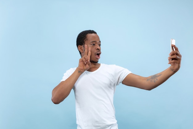 Half-length close up portrait of young african-american man in white shirt on blue space. Human emotions, facial expression, ad concept