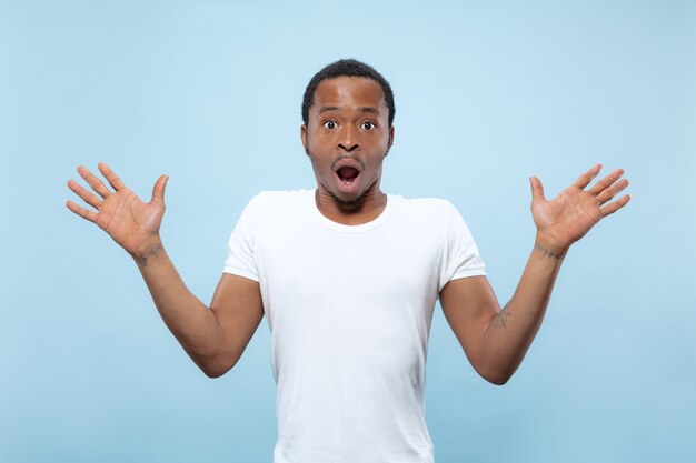 Half-length close up portrait of young african-american man in white shirt on blue space. Human emotions, facial expression, ad, sales concept. Inviting, looks shocked and astonished