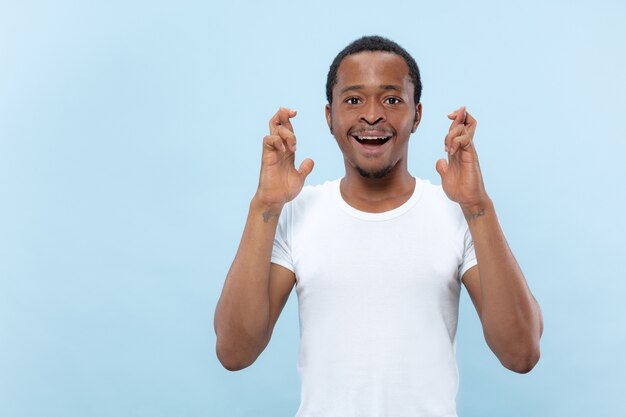 Half-length close up portrait of young african-american man in white shirt on blue wall. Human emotions, facial expression, ad concept. Showing the sign of good luck, smiling, hopeful.