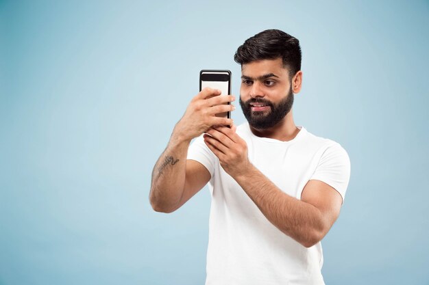 Half-length close up portrait of young hindoo man in white shirt on blue background. Human emotions, facial expression, ad concept. Negative space. Making selfie or videoblog, vlog, chating.