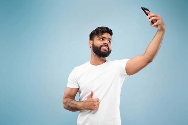 Half-length close up portrait of young hindoo man in white shirt on blue background. Human emotions, facial expression, ad concept. Negative space. Making selfie or videoblog, vlog, chating.