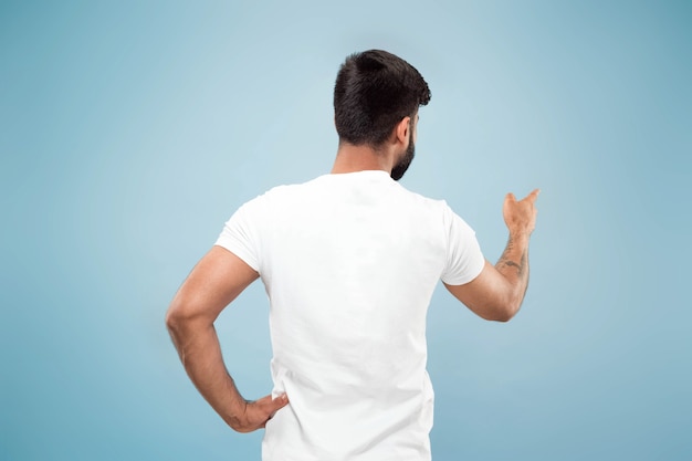 Half-length close up portrait of young hindoo man in white shirt on blue background. Human emotions, facial expression, ad concept. Negative space. Showing empty bar, pointing, choosing, inviting.