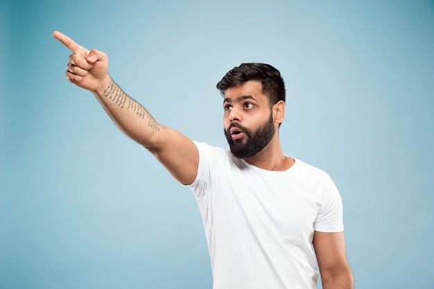 Half-length close up portrait of young hindoo man in white shirt on blue background. Human emotions, facial expression, sales, ad concept. Negative space. Pointing up being shocked and astonished.