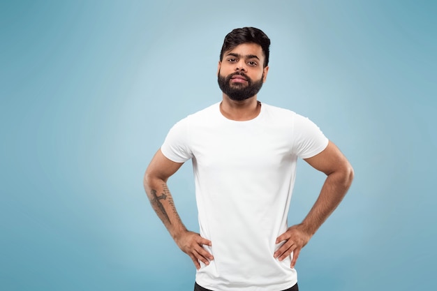 Half-length close up portrait of young hindoo man in white shirt on blue wall. Human emotions, facial expression, ad concept. Negative space. Posing, standing and smiling, looks calm.