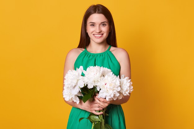 Half length of lady in elegant green dress keeps bouquet of flowers in hands on yellow, being happy to recive peonies as presenmt.