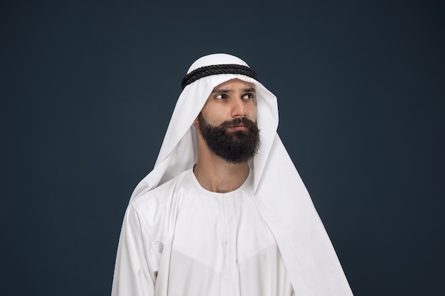 Free photo half-length portrait of arabian saudi businessman on dark blue  space. young male model standing and looks thoughtful