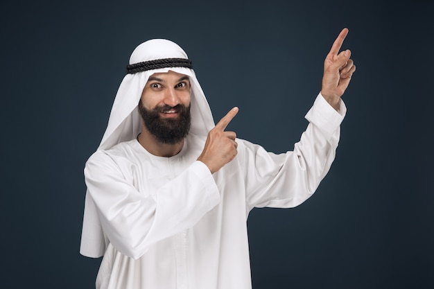 Half-length portrait of arabian saudi businessman on dark blue studio background. Young male model smiling and pointing or choosing. Concept of business, finance, facial expression, human emotions.