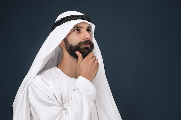 Half-length portrait of arabian saudi businessman on dark blue studio background. Young male model standing and looks thoughtful. Concept of business, finance, facial expression, human emotions.