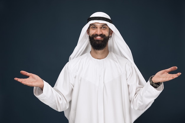 Free Photo half-length portrait of arabian saudi businessman on dark blue wall. young male model smiling, showing a gesture of inviting. concept of business, finance, facial expression, human emotions.