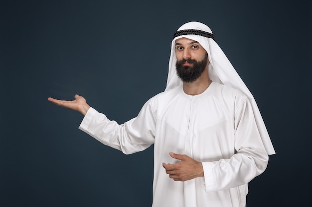 Half-length portrait of arabian saudi man on dark blue  space. Young male model smiling and pointing