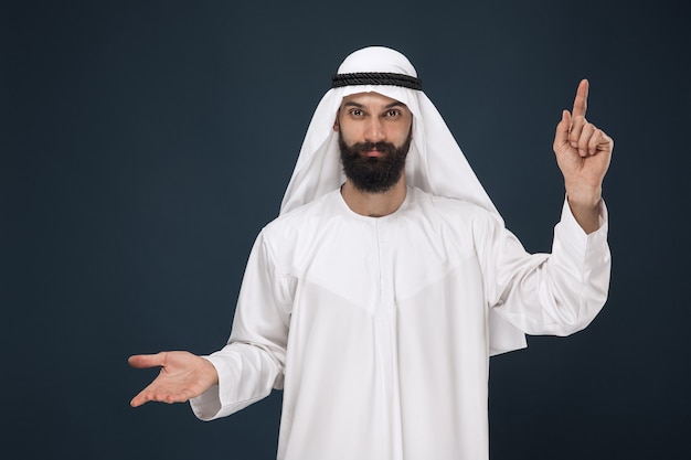 Half-length portrait of arabian saudi man on dark blue studio background. Young male model smiling and pointing. Concept of business, finance, facial expression, human emotions, technologies.