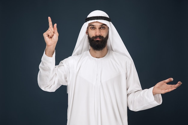 Half-length portrait of arabian saudi man on dark blue wall. Young male model smiling and pointing. Concept of business, finance, facial expression, human emotions, technologies.