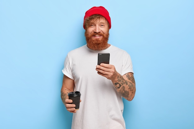 Half length shot of cheerful bearded redhead man wears stylish hat, white casual t shirt, holds mobile phone, takeout coffee, being in good mood, types messages