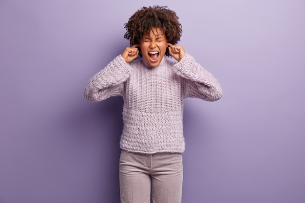 Free photo half length shot of depressed mixed race woman plugs ears, doesnt want to hear quarrel, annoyed of gossips, wears knitted jumper and trousers, stands over purple  wall. ignoring noise