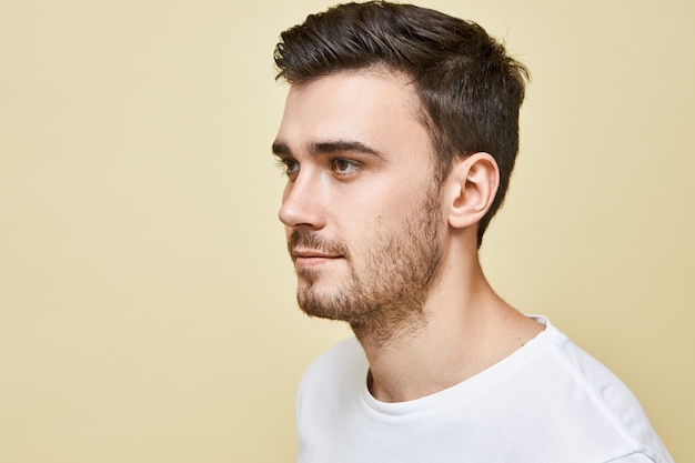 Half profile image of handsome young Caucasian man with good skin, brown eyes, black stylish hair and stubble posing isolated against blank wall, looking in front of him, smiling