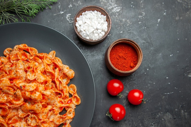 Half shot of easy pasta meal for dinner on a black plate and different spices and tomatoes on a dark background