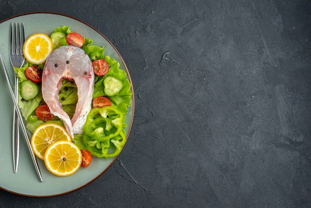 Free photo half shot of raw fish and fresh vegetables lemon slices and cutlery set on a gray plate on the left side on black surface with free space