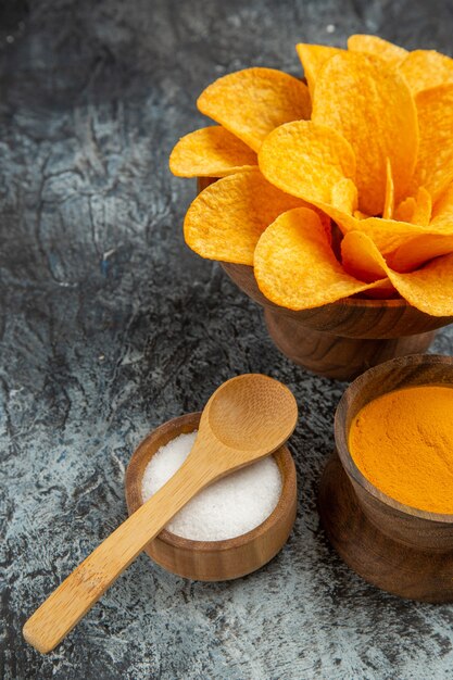 Half shot of tasty potato chips decorated like flower shaped different spices with spoons on them on the left side of gray table