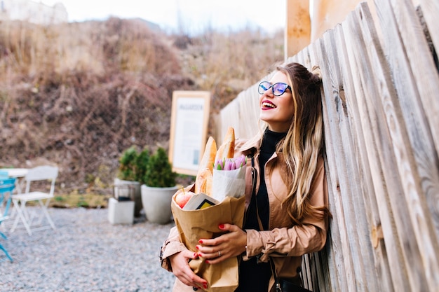 Half-turned photo of attractive woman with paper bag of tasty meal from supermarket. Charming girl carrying fresh foodstuffs for lunch with her family posing with dreamy face expression.