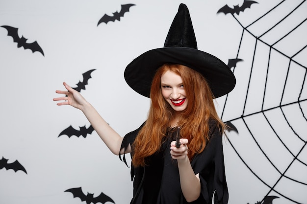 Halloween Concept - Beautiful Witch playing with magic stick on grey wall.