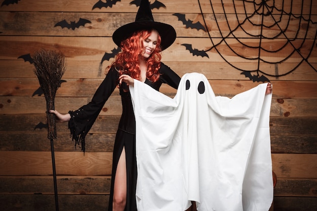 Free Photo halloween concept - witch mother and little white ghost doing trick or treat celebrating halloween posing with curved pumpkins over bats and spider web on wooden studio background.