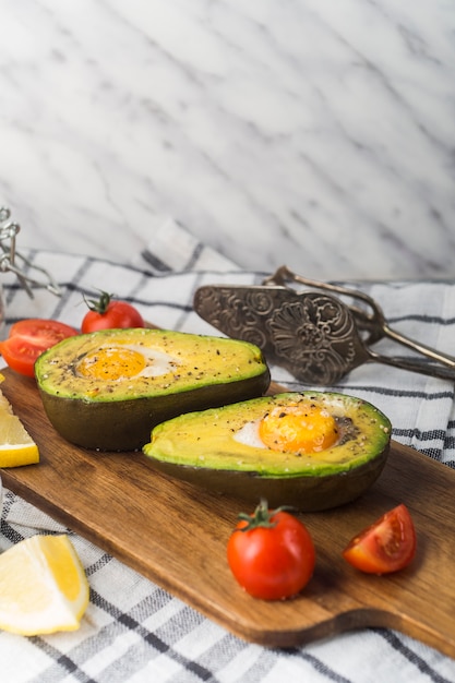 Free Photo halved avocado with egg york; tomatoes and lemon slice on chopping board over the napkin with antique tong