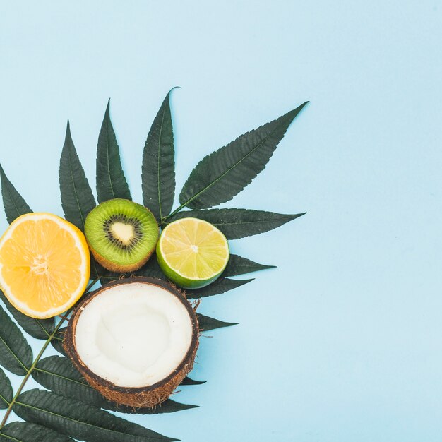 Halved coconut; lemon; orange and kiwi on green leaves against blue background