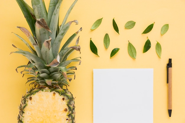 Free photo halved pineapple with leaves; pen and white paper on yellow background