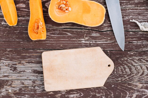 Halves of raw organic butternut squash with chopping board and sharp knife on weathered table top