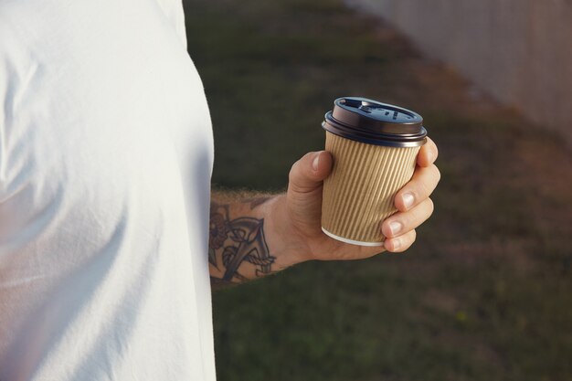 hand and chest of a white tattooed man wearing white unlabeled t-shirt holding a light brown paper coffee cup