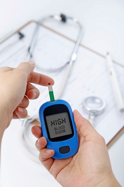 Hand holding a blood glucose meter measuring blood sugar, the background is a stethoscope and chart file