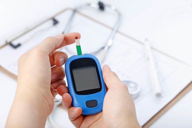 Hand holding a blood glucose meter measuring blood sugar, the background is a stethoscope and chart file