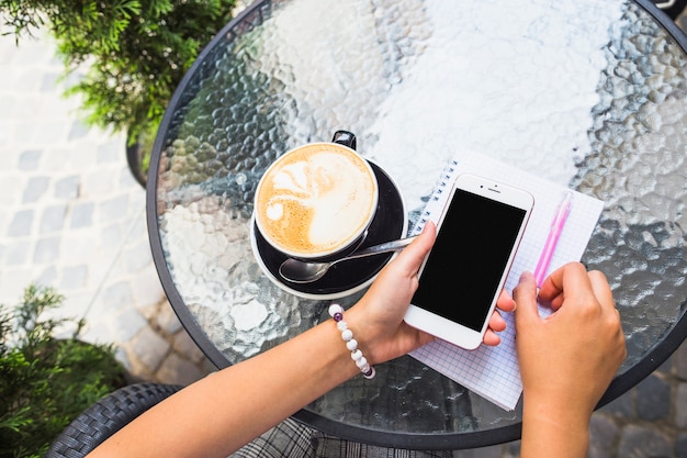 Hand holding cellphone with coffee cup on glass table