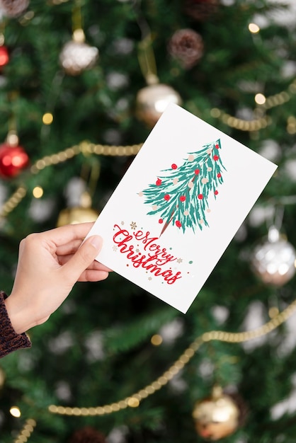 Free photo hand holding christmas greeting card on christmas tree background.