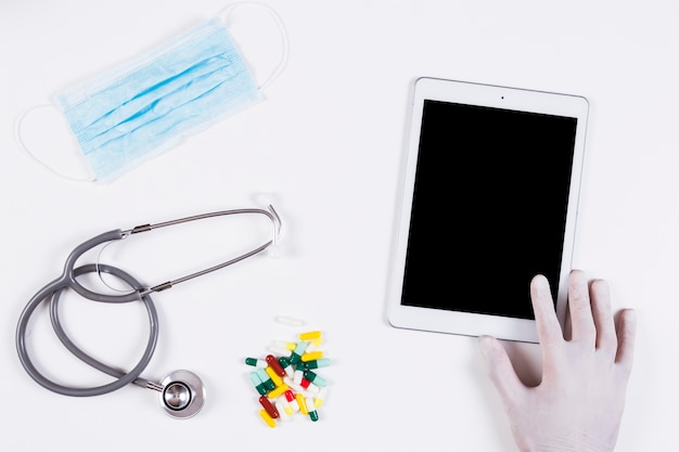 Free Photo hand holding digital tablet with mask; stethoscope and colorful capsules on white background