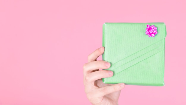 Free Photo hand holding green wrapped gift box with pink bow against colored background