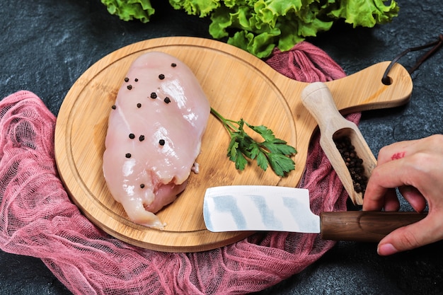 Hand holding knife and piece of raw chicken fillet on wooden plate with fresh vegetables and tablecloth.