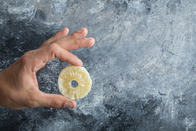 Free photo hand holding a slice of dried pineapple on a gray background.