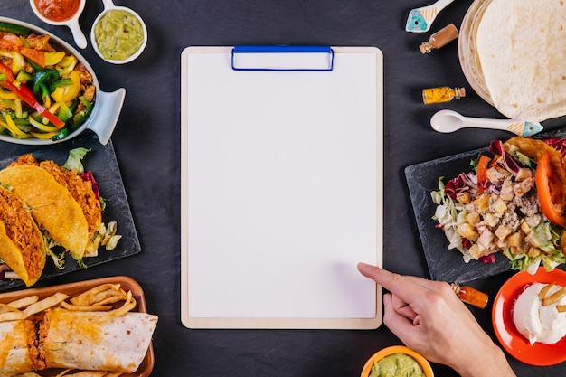 Free Photo hand pointing at clipboard among mexican food